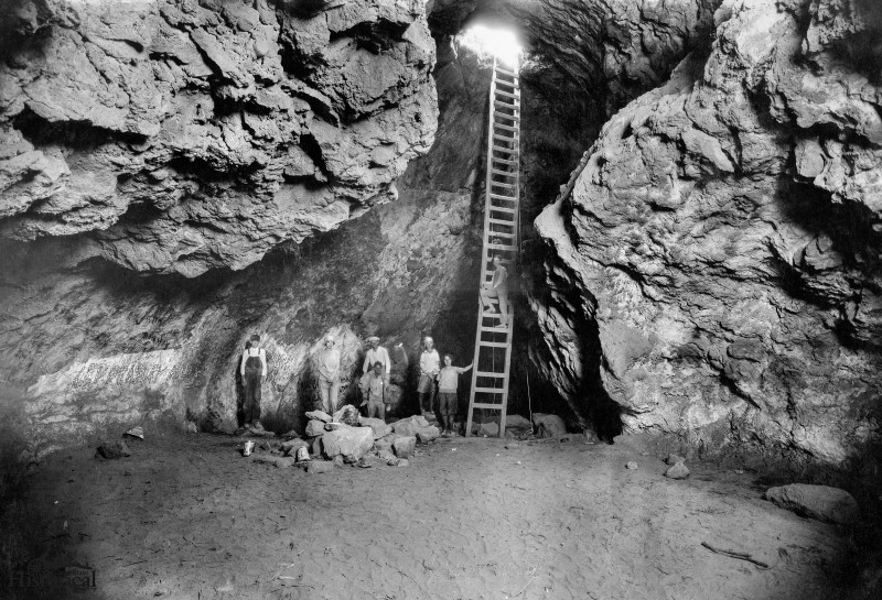 "decorative image: old black and white tintype image of a group of miners in a cave - one is climbing up a ladder towards the light"